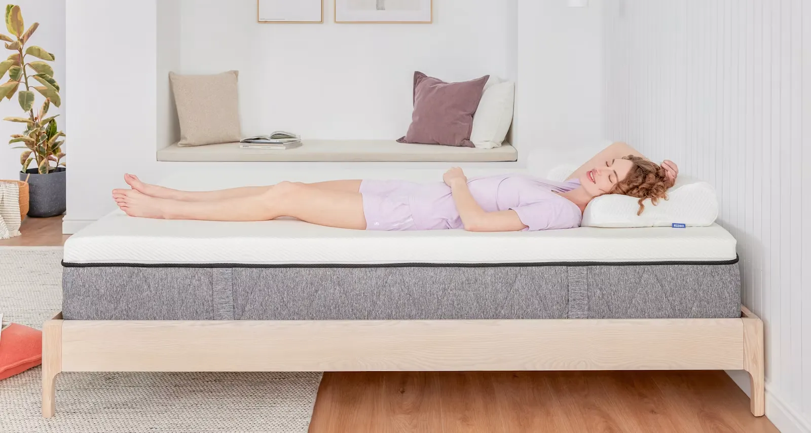 A woman lying on her back on the Ecosa mattress, showing its white quilted top and grey knit side panel on a low timber bed base.