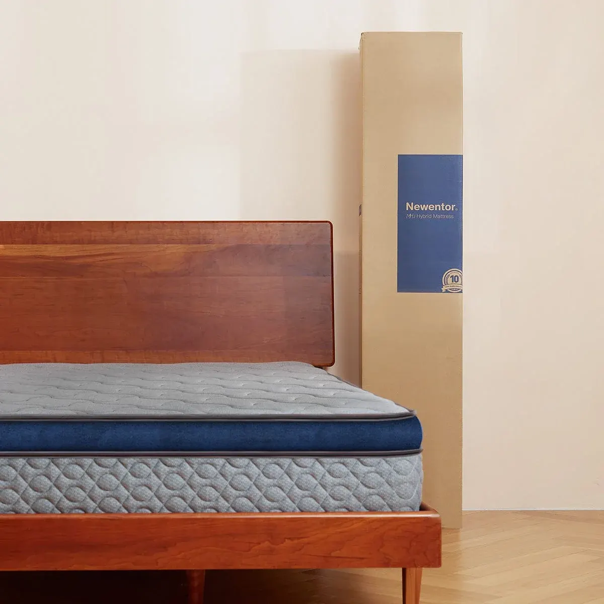 The Newentor MH2 Hybrid mattress on a timber bed frame with its delivery box standing alongside, showing the grey quilted top and blue side band.