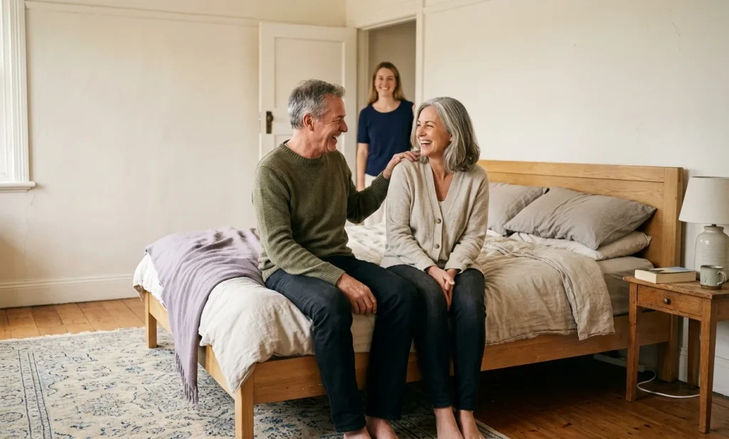 An older couple sit laughing together on the edge of a well-used oak bed with soft linen bedding, while their adult daughter watches from the doorway behind them.