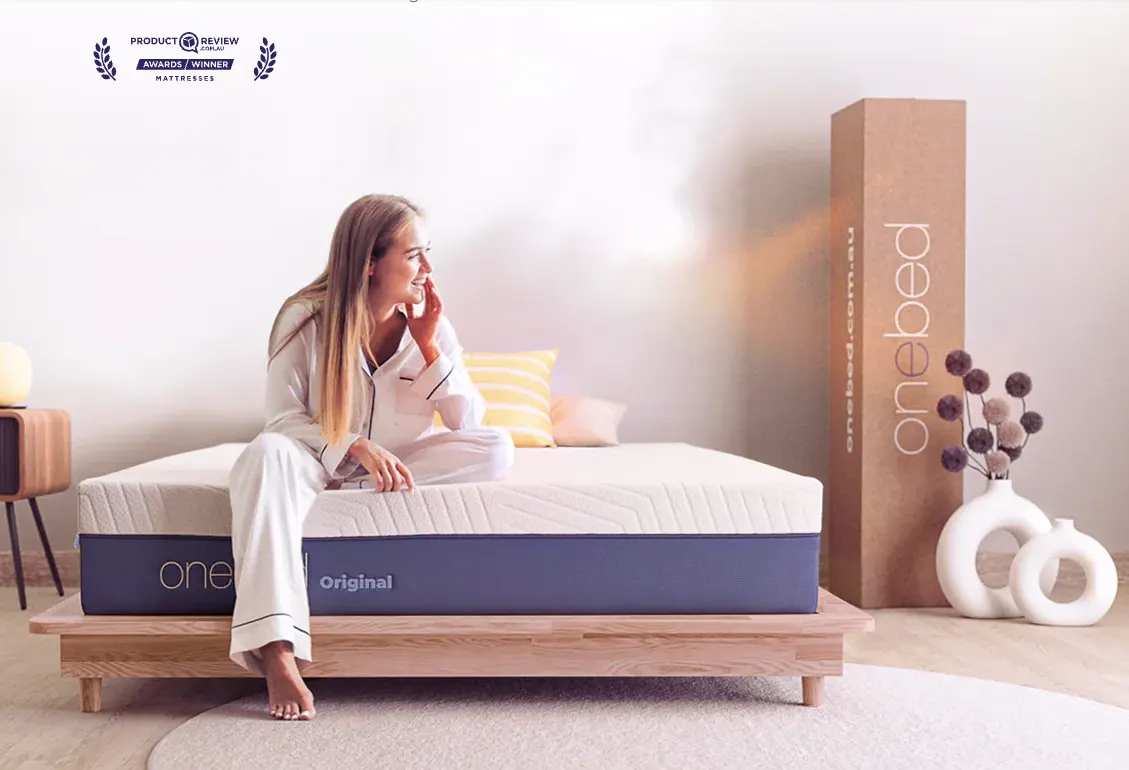 A woman sitting on the edge of the Onebed Original mattress, showing its cream top and navy blue side panel, with the delivery box alongside.